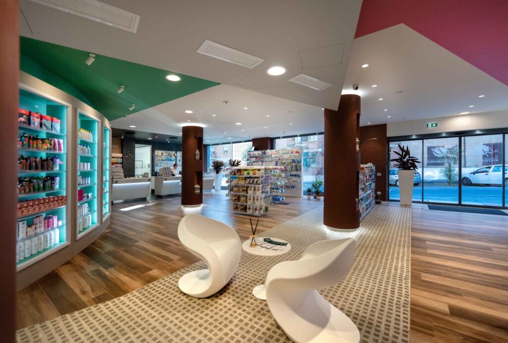 Vista ampia farmacia con area attesa, espositori centrali e ingresso vetrato con luce naturale