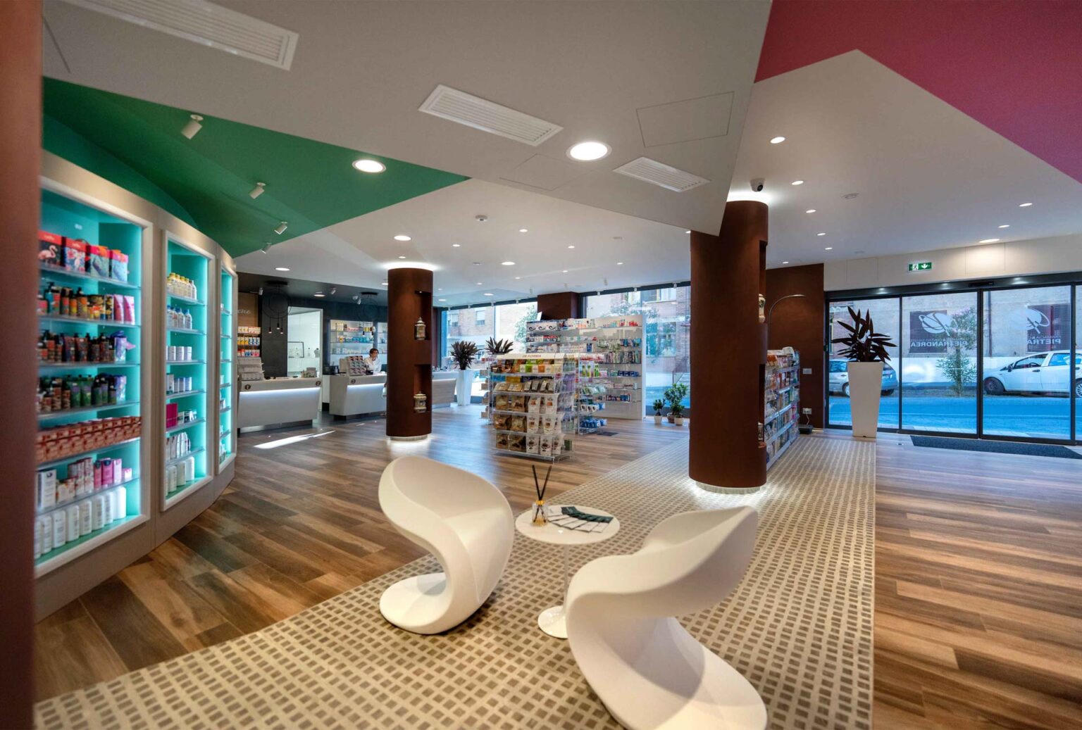 Vista ampia farmacia con area attesa, espositori centrali e ingresso vetrato con luce naturale